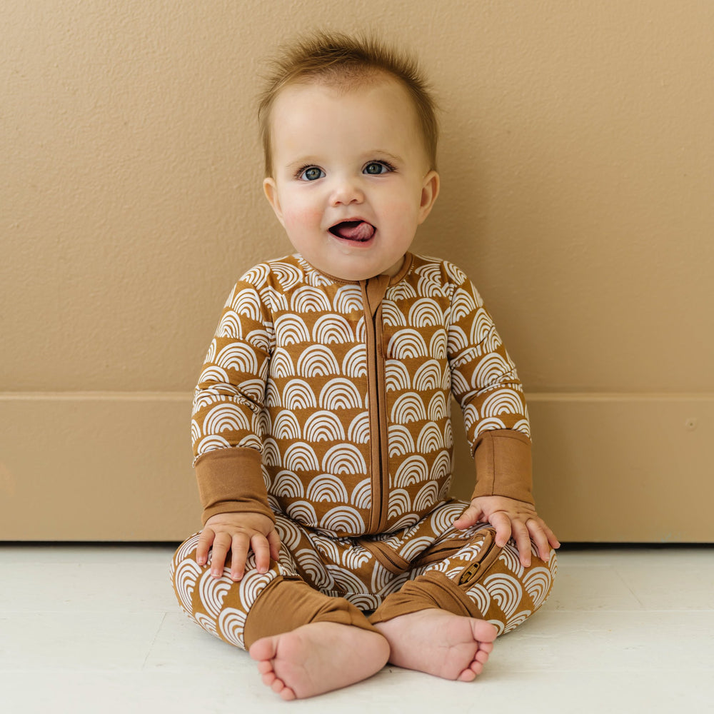 Image of infant baby wearing a long sleeve zip up romper in Rust Rainbows print. This print features white rainbows that sit upon a rust brown background with matching rust brown trim.