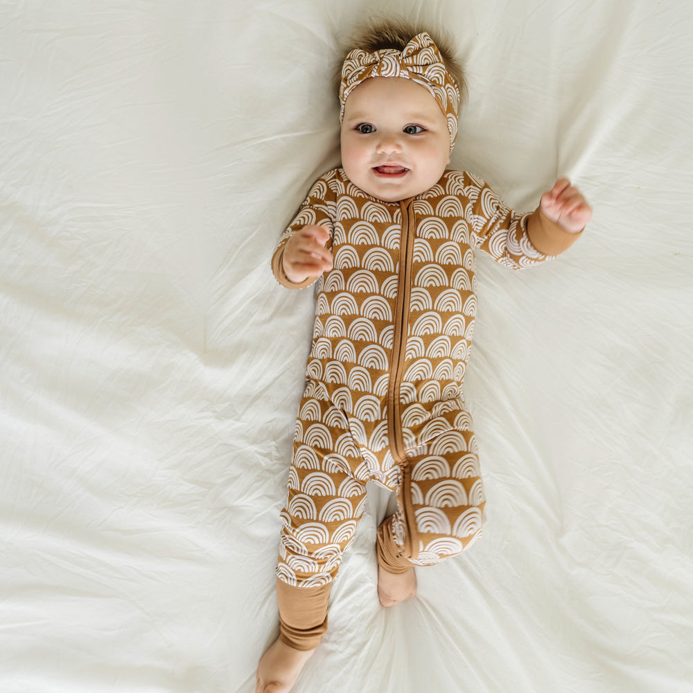 Image of infant baby wearing a long sleeve zip up romper and coordinating bow headband in Rust Rainbows print. This print features white rainbows that sit upon a rust brown background with matching rust brown trim.