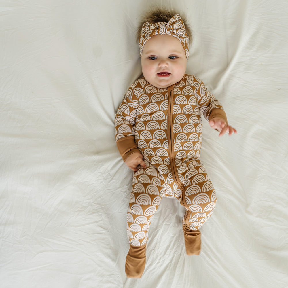 Image of infant baby wearing a long sleeve zip up romper and coordinating bow headband in Rust Rainbows print. This print features white rainbows that sit upon a rust brown background with matching rust brown trim.