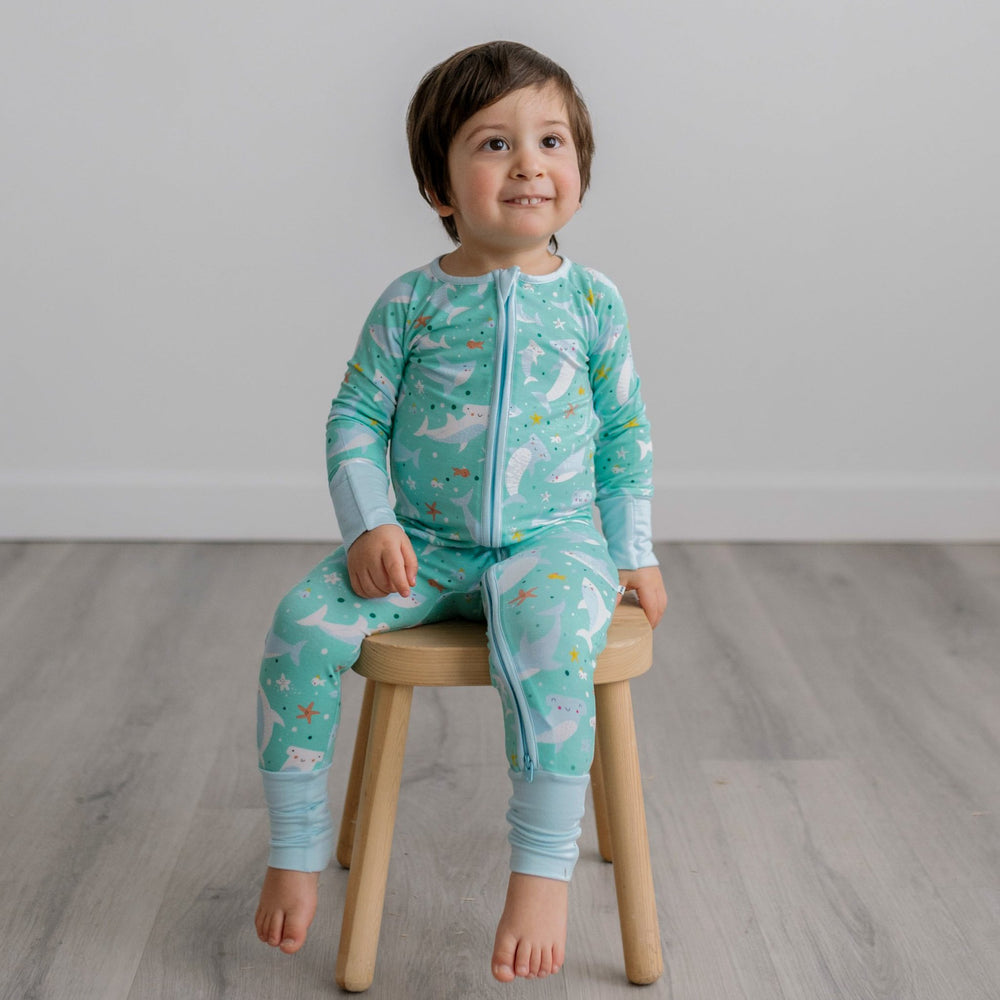 Image of toddler boy sitting on a wooden stool. He is shown wearing a shark printed zip up romper. This print includes hammerhead and great white sharks, featured on an aqua background with a light blue trim. 