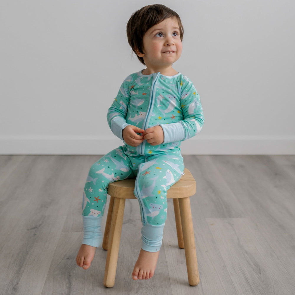 Image of toddler boy sitting on a wooden stool. He is shown wearing a shark printed zip up romper. This print includes hammerhead and great white sharks, featured on an aqua background with a light blue trim.