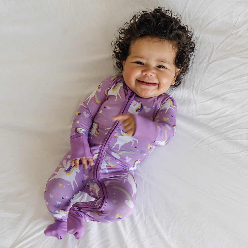 Image of infant girl wearing a unicorn printed zip up romper. Flying unicorns with rainbow-colored manes gallop across a purple background with hearts, stars, and rainbows in this magical print.