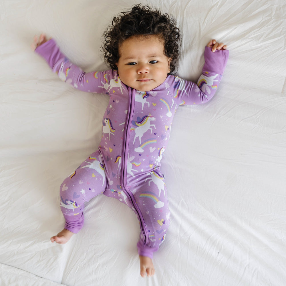 Click to see full screen - Image of infant girl wearing a unicorn printed zip up romper. Flying unicorns with rainbow-colored manes gallop across a purple background with hearts, stars, and rainbows in this magical print.